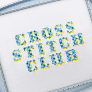 May include: A white fabric stretched within a gray embroidery hoop, featuring the words "CROSS STITCH CLUB" in blue cross-stitch letters with yellow outlines. The hoop is marked with "150x150".