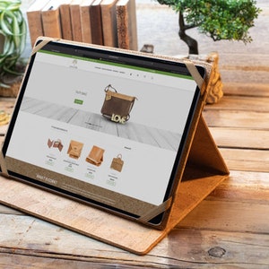 May include: A brown leather tablet case with a stand, displaying a website on a tablet screen. The website features a variety of leather bags and purses, including a brown crossbody bag with a gold "LOVE" charm.