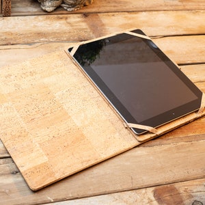 Puede incluir: Una funda rectangular para tableta hecha de material de corcho de color marr&oacute;n claro con motas. La funda presenta un dise&ntilde;o plano tipo libro abierto con correas el&aacute;sticas en las esquinas que aseguran una tableta negra, mostrando una textura natural.
