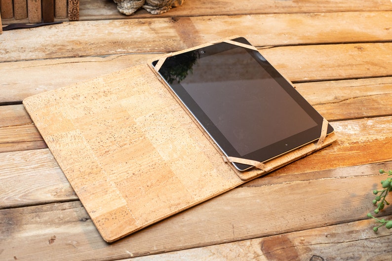 Puede incluir: Una funda rectangular para tableta hecha de material de corcho de color marr&oacute;n claro con motas. La funda presenta un dise&ntilde;o plano tipo libro abierto con correas el&aacute;sticas en las esquinas que aseguran una tableta negra, mostrando una textura natural.