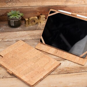May include: A cork iPad case with a brown cork exterior and a black screen. The case is open and the iPad is visible. A white stylus is resting on the case.