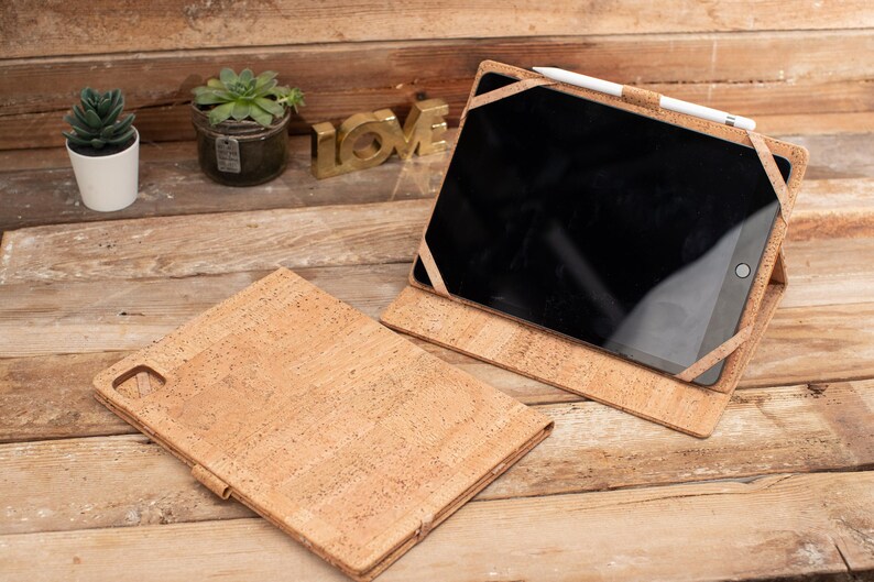 May include: A cork iPad case with a brown cork exterior and a black screen. The case is open and the iPad is visible. A white stylus is resting on the case.