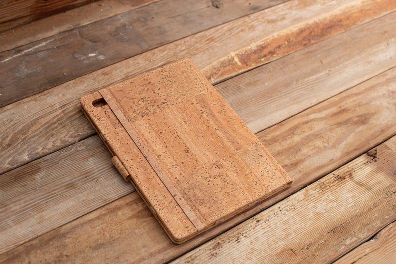 Puede incluir: Una funda rectangular para tableta hecha de material de corcho natural con una textura moteada en tonos bronceado y marr&oacute;n. La funda cuenta con una correa el&aacute;stica vertical en la cubierta frontal y una peque&ntilde;a abertura en la esquina.