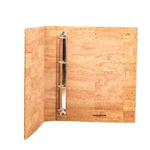 May include: A brown cork binder with a silver three-ring mechanism. The binder is open to reveal the inside, which is also made of cork. The binder has the word "TAMPONCINE" printed on the front cover.