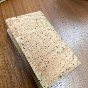 May include: A small, light brown cork wallet with a stitched edge. The wallet is made of natural cork and has a subtle, speckled pattern.