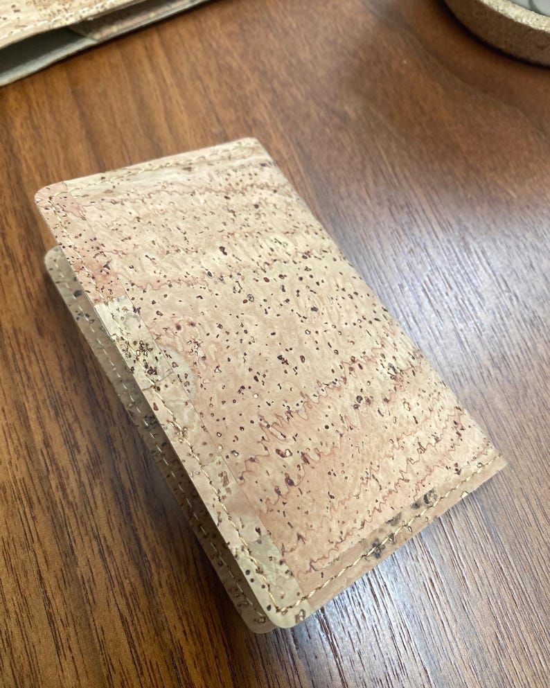 May include: A small, light brown cork wallet with a stitched edge. The wallet is made of natural cork and has a subtle, speckled pattern.