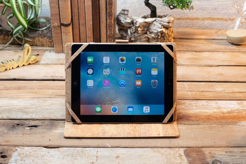 Puede incluir: Una tableta asegurada en una funda protectora de color marr&oacute;n claro con textura de corcho y correas el&aacute;sticas en las esquinas. La pantalla muestra una cuadr&iacute;cula est&aacute;ndar de iconos de aplicaciones, incluyendo mensajes, calendario, fotos, c&aacute;mara y ajustes.
