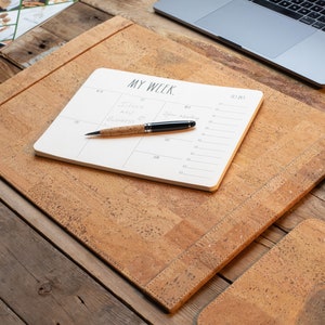 May include: A cork desk setup featuring a desk mat, a notepad with the text "MY WEEK," and a pen. The desk mat and pen have a natural cork finish. A laptop and mouse are also visible on the wooden desk.