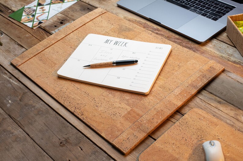 May include: A cork desk setup featuring a desk mat, a notepad with the text "MY WEEK," and a pen. The desk mat and pen have a natural cork finish. A laptop and mouse are also visible on the wooden desk.