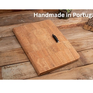 Può includere: Una cartella rettangolare in sughero marrone chiaro con una superficie testurizzata e una penna nera inserita in una piccola fessura sulla copertina anteriore. Il testo Handmade in Portugal è stampato in lettere bianche in alto.