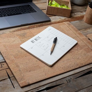 May include: A cork desk set featuring a 48 x 33 cm desk mat, a mouse pad, and a pen holder. A laptop, a notepad with the text "MY WEEK", and a pen are on the desk. The desk is made of wood.