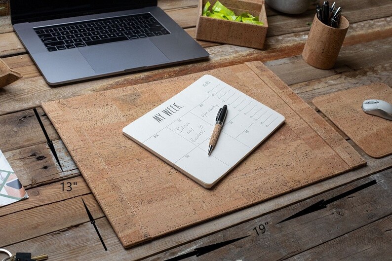 May include: A cork desk set featuring a 48 x 33 cm desk mat, a mouse pad, and a pen holder. A laptop, a notepad with the text "MY WEEK", and a pen are on the desk. The desk is made of wood.