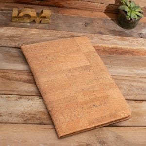 May include: A light brown cork notebook with a textured surface. The notebook is lying on a wooden surface.