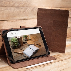 Peut inclure: Une housse en cuir marron pour iPad avec un extérieur en liège. La housse est pliée pour maintenir l'iPad en position verticale, qui affiche un site Web avec le texte "A choice you can feel good about". Un stylet Apple blanc est posé sur la table à côté de la housse.