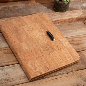 May include: A cork notebook with a black pen on top of it. The notebook is lying on a wooden surface.