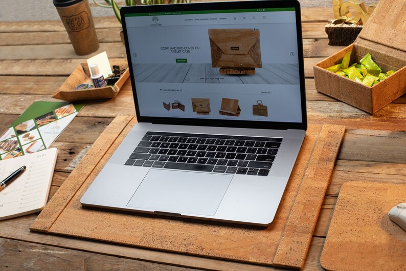 May include: A laptop on a cork desk mat, with a matching mouse mat and a coffee cup. The laptop screen shows a website selling cork products, including a tablet case. The desk is made of wood.