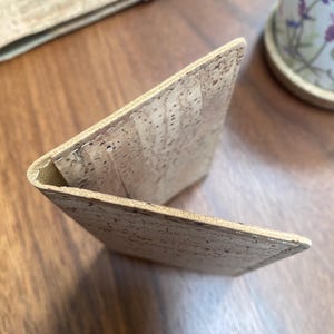 May include: A light brown cork wallet folded in half, showing the inside of the wallet. The wallet is made of a natural cork material with a textured surface.