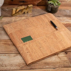 May include: A square, light brown cork portfolio folder featuring a textured, natural speckled surface. The folder includes a horizontal pocket containing a small dark green card and a black pen resting diagonally across the top edge of the pocket.