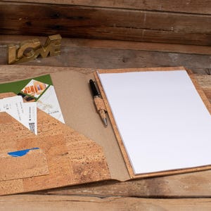 May include: A cork-covered folder with a white notepad and a black pen. The folder has a pocket on the left side with a green folder, a blue card, and two tickets inside.