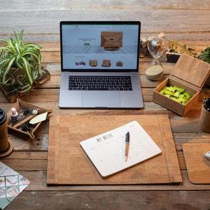 May include: A home office setup with a laptop showing a website, a cork desk pad, a mouse pad, and a notebook with the text "MY WEEK". Other items include a coffee mug, a pen holder, and a small box. Plants and an hourglass are also present.