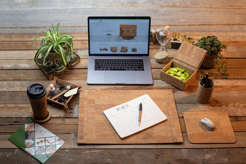 May include: A home office setup with a laptop showing a website, a cork desk pad, a mouse pad, and a notebook with the text "MY WEEK". Other items include a coffee mug, a pen holder, and a small box. Plants and an hourglass are also present.