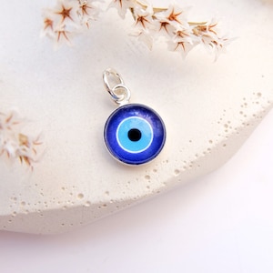 Puede incluir: Un amuleto de plata con un amuleto de ojo azul. El amuleto es redondo y tiene un dise&ntilde;o de ojo blanco y azul.