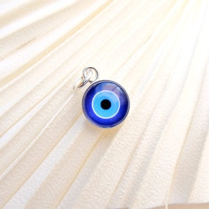 Puede incluir: Un amuleto de plata con un dise&ntilde;o de ojo azul malvado. El amuleto es redondo y tiene un c&iacute;rculo blanco en el centro con una pupila negra.