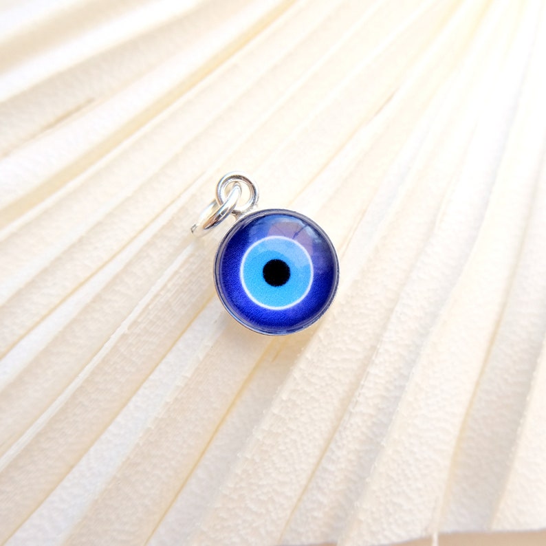 Puede incluir: Un amuleto de plata con un dise&ntilde;o de ojo azul malvado. El amuleto es redondo y tiene un c&iacute;rculo blanco en el centro con una pupila negra.