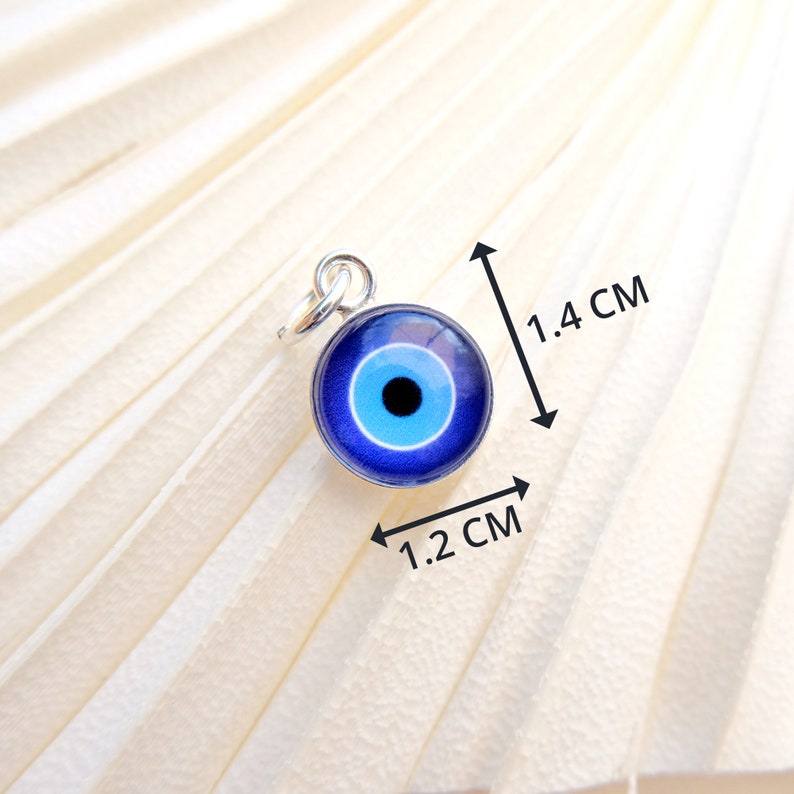 Puede incluir: Un amuleto de plata con un dise&ntilde;o de ojo azul malvado. El amuleto mide 1,2 cm de ancho y 1,4 cm de alto.