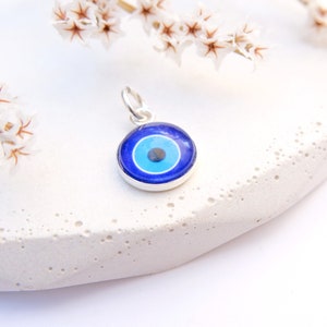 Puede incluir: Un amuleto de plata con un dise&ntilde;o de ojo azul malvado. El amuleto es redondo y tiene un fondo blanco con un ojo azul en el centro. El ojo est&aacute; rodeado por un c&iacute;rculo blanco.
