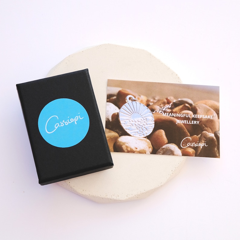Puede incluir: Una caja negra con un c&iacute;rculo azul que dice "Cassiopi". Una tarjeta blanca con el texto "Meaningful Keepsake Jewellery" y "Cassiopi" en ella. La tarjeta est&aacute; al lado de un colgante de plata con un dise&ntilde;o de sol y olas.