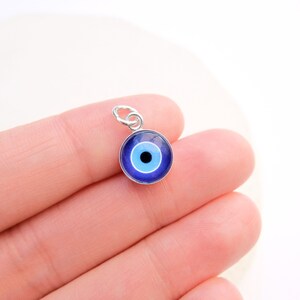Puede incluir: Un amuleto de plata con un dise&ntilde;o de ojo azul malvado. El ojo est&aacute; rodeado por un anillo blanco y una pupila negra.