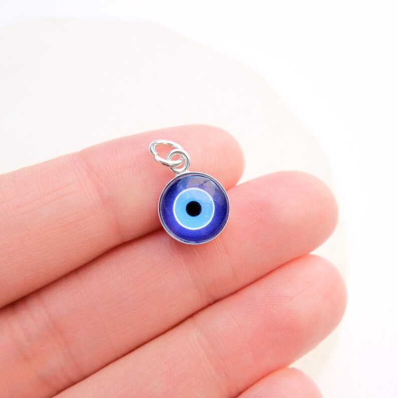 Puede incluir: Un amuleto de plata con un dise&ntilde;o de ojo azul malvado. El ojo est&aacute; rodeado por un anillo blanco y una pupila negra.