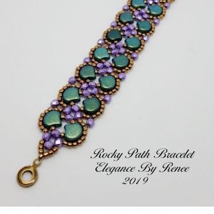 May include: A beaded bracelet with a pattern of green and purple beads. The bracelet has a gold clasp and is labelled "Rocky Path Bracelet Elegance By Renee 2019".