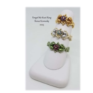 Pode incluir: Três anéis de contas com desenhos de flores. Os anéis são dourados, prateados e verdes com flores roxas, brancas e vermelhas.