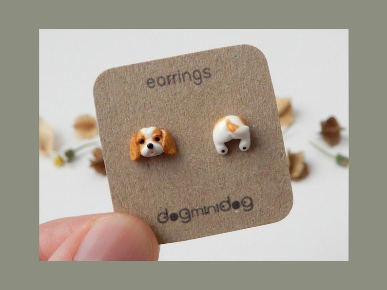 May include: A pair of earrings featuring a small, white and brown ceramic dog head on one side and a small, white and brown ceramic dog tail on the other side. The earrings are displayed on a brown card with the text "earrings" and "dogminidog".