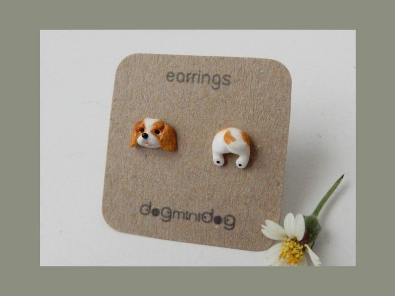 May include: A pair of earrings featuring a Cavalier King Charles Spaniel dog head and a Cavalier King Charles Spaniel dog bottom. The earrings are on a brown card with the text "earrings" and "dognidoG".