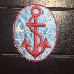 May include: A red embroidered anchor with white waves and a sailboat on a blue oval background.