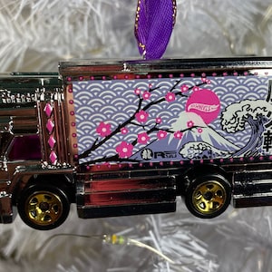 May include: A silver Hot Wheels truck ornament with a Japanese-inspired design. The truck features a pink cherry blossom branch, a blue and white wave, and Mount Fuji. The truck also has the text "Hot Wheels" and "風火輪" on the side.