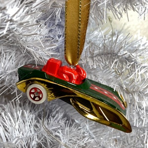 May include: A green and gold Hot Wheels car ornament with a red figure riding in the car. The ornament is hanging from a gold ribbon on a white artificial Christmas tree.