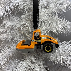 May include: An orange and black Matchbox road roller ornament with a black ribbon for hanging. The ornament is set against a white Christmas tree background. The road roller has a gray roller and black wheels.