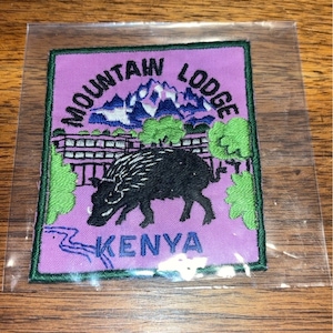May include: A purple embroidered patch with green trim. The patch features a black wild boar, a mountain range, and a building. The text "Mountain Lodge Kenya" is embroidered in blue.