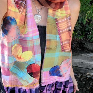 May include: A patchwork scarf with a variety of plaid patterns in pink, orange, yellow, and green. The scarf features heart-shaped appliqués and a ruffled hem. A silver heart pendant hangs from a chain.