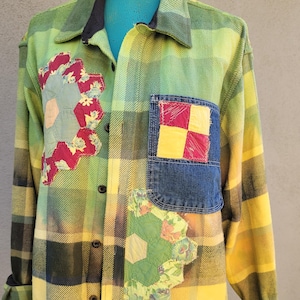 May include: A green and yellow plaid flannel shirt with patchwork details. The shirt features floral and geometric patches in shades of red, yellow, and green. The shirt has a denim pocket and button-down front.