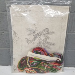May include: A needlepoint kit featuring a bird on a branch with berries. The kit includes a piece of Aida cloth, embroidery floss, and instructions. The kit is packaged in a clear plastic bag.