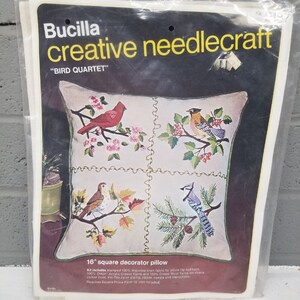 May include: A Bucilla creative needlecraft kit for a 16-inch square pillow featuring a "Bird Quartet" design. The pillow is divided into four sections, each depicting a different bird: a cardinal, a robin, a sparrow, and a blue jay. The kit includes stamped linen fabric, acrylic and wool yarns, cotton floss, piping, a zipper, a needle, and instructions.