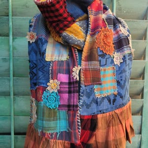 May include: A patchwork scarf with a variety of fabrics in red, blue, yellow, and orange. The scarf has a ruffled hem and is adorned with crocheted floral accents. The design incorporates plaid, solid, and patterned fabrics.