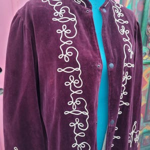 May include: A long, dark purple velvet jacket with white embroidered details. The jacket has a mandarin collar and a button closure.