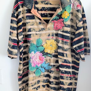 May include: A short-sleeve polo shirt featuring a horizontal striped pattern in navy blue, tan, and red. The fabric has a bleached, mottled effect throughout. A large floral graphic in pink, yellow, and teal is printed on the front and shoulder.