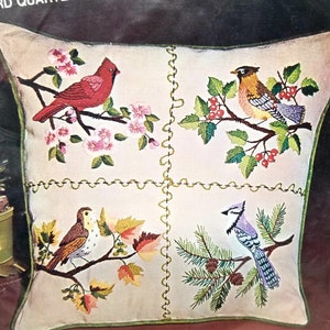 May include: A white pillow with four embroidered squares featuring different birds and foliage. The top left square shows a red cardinal on a branch with pink blossoms. The top right square shows a robin on a branch with green leaves and red berries. The bottom left square shows a brown bird on a branch with fall leaves. The bottom right square shows a blue jay on a branch with pine needles and pine cones.